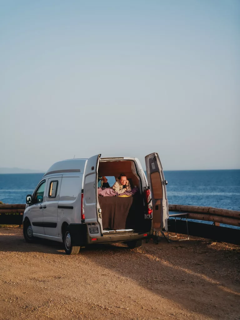 Vanlife en Sardaigne : van avec les portes arrière ouverte et une femme qui regarde la vue sur la mer.