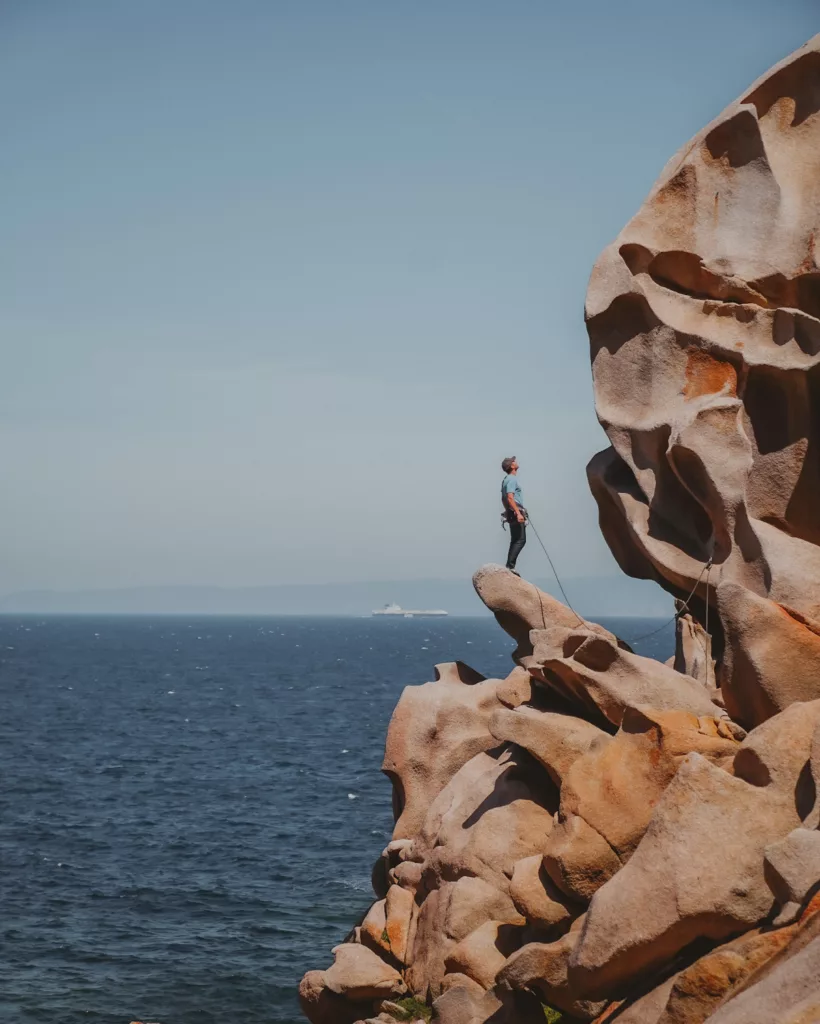 Grimpeur face à la falaise d'escalade de granit arrondie de Capo testa avec la mer et un navire en arrière-plan.